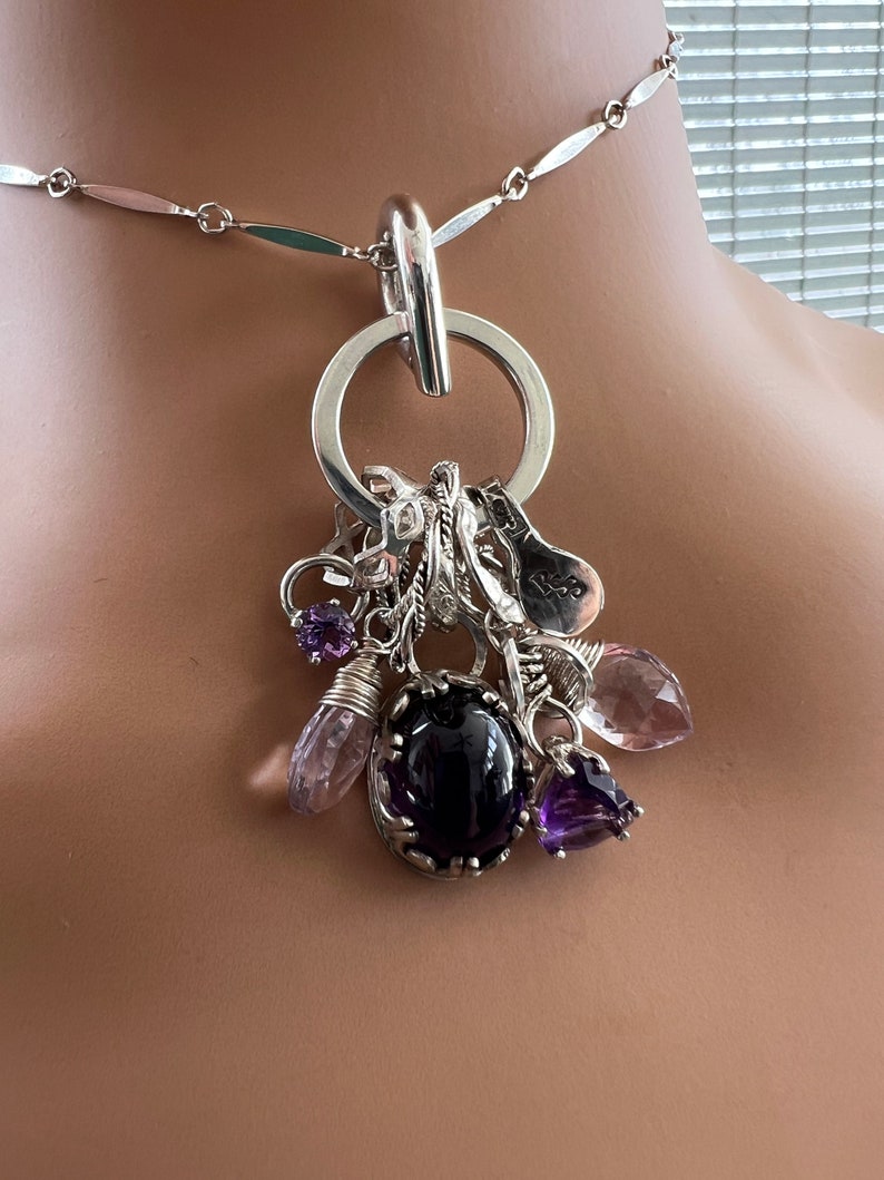 Puede incluir: Collar de plata con un gran colgante circular y un conjunto de abalorios. Los abalorios incluyen varias piedras preciosas moradas y peque&ntilde;os adornos plateados. El collar tiene una cadena delicada con eslabones alargados.