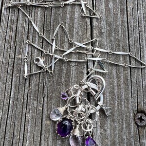 Puede incluir: Un collar de plata con una cadena delicada y un grupo de piedras preciosas. Las piedras preciosas incluyen amatista y cuarzo transparente en varias formas. El collar se muestra sobre una superficie de madera envejecida.