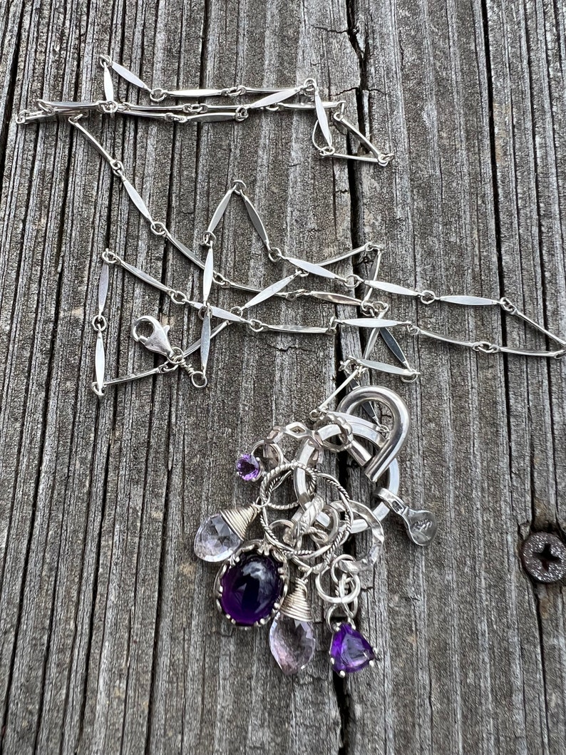 Puede incluir: Un collar de plata con una cadena delicada y un grupo de piedras preciosas. Las piedras preciosas incluyen amatista y cuarzo transparente en varias formas. El collar se muestra sobre una superficie de madera envejecida.