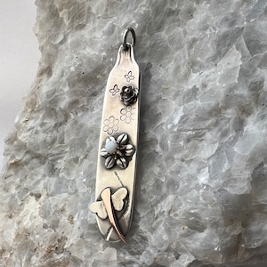 May include: A silver spoon pendant with a dragonfly design, flowers, and a white opal stone. The pendant is on a white rock background.