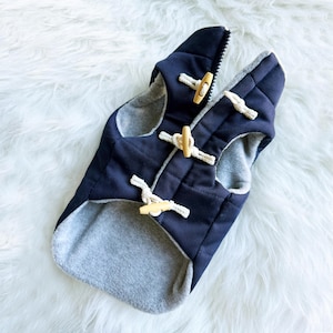 May include: Navy blue dog coat with a quilted exterior and a gray fleece lining. The coat features wooden toggle closures and a zipper. The dog coat is designed to keep your pet warm and comfortable.