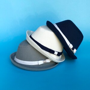 May include: Three hats stacked on top of each other. The top hat is navy blue with a white band. The middle hat is white with a black band. The bottom hat is gray with a white band.