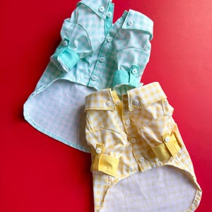 May include: Two dog shirts in a gingham pattern. One is light blue and white, the other yellow and white. Both have buttoned cuffs and collars. The shirts are on a red background.