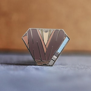 May include: A diamond-shaped enamel pin with a brown and tan colour scheme, featuring a stylised "V" design. The pin has a metallic border and a light blue accent. The text "ARMSTRONG OUTPOST" is at the bottom.