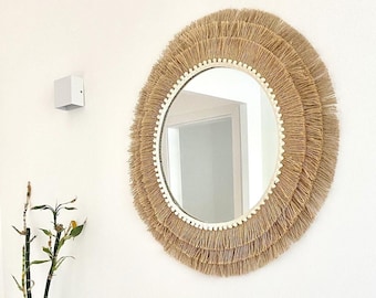 May include: A round, decorative mirror with a natural fiber fringe. The mirror has a gold-colored frame and is mounted on a white wall. Below the mirror is a white shelf with candles, a black tray, and decorative objects.