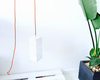 May include: A white rectangular pendant light fixture hangs from a bright orange cord against a white wall. The light fixture is made of a textured material and is suspended from the ceiling.