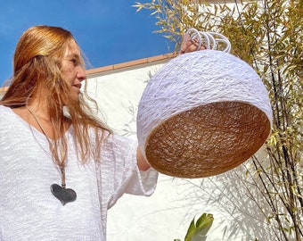 May include: A person holds a large, white, round, woven lampshade with a brown inner lining. The lampshade is made of natural fibers and has a rustic, bohemian style.
