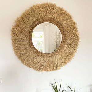 May include: Round wall mirror with a natural woven raffia frame. The mirror has a circular glass surface and a wide, fringed border. The frame is a light tan color, and the mirror is hanging on a white wall. The overall style is bohemian.