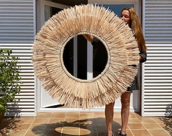 May include: A large, round, woven wall hanging made of natural materials. The hanging is in the shape of a sun with a large, open center. The hanging is being held by a person in front of a brick wall.