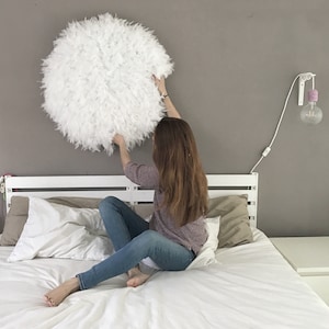May include: A white feather wall hanging is being hung on a white wall above a white bed with white bedding and a brown pillow. A person is hanging the wall hanging.