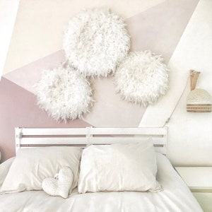 Peut inclure: Un lit blanc avec une tête de lit blanche et une literie blanche. Il y a trois décorations murales rondes et moelleuses blanches sur le mur derrière le lit. Le mur est peint dans des tons de rose et de blanc.