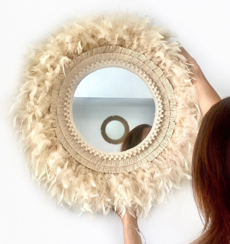 May include: A round mirror with a cream-coloured woven frame and a fringe of feathers. The mirror is hanging on a white wall.
