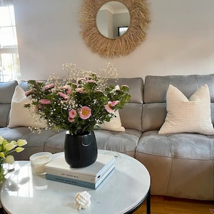 May include: A grey sofa with white cushions and a bouquet of pink and white flowers in a black vase. The flowers are on a white marble coffee table with two books and a white rope knot. The wall behind the sofa is white with a large woven mirror.