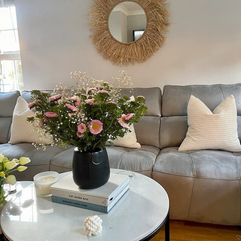 May include: A grey sofa with white cushions and a bouquet of pink and white flowers in a black vase. The flowers are on a white marble coffee table with two books and a white rope knot. The wall behind the sofa is white with a large woven mirror.