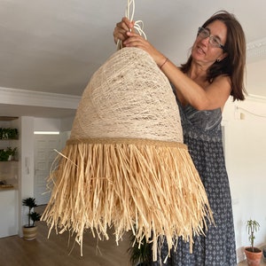 K&ouml;nnte beinhalten: Ein gro&szlig;er, runder, geflochtener Lampenschirm mit einem nat&uuml;rlichen, stroh&auml;hnlichen Fransenbesatz. Der Lampenschirm h&auml;ngt an einem wei&szlig;en Kabel und wird von einer Person gehalten.