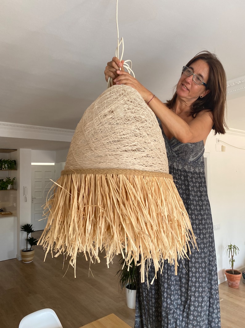 K&ouml;nnte beinhalten: Ein gro&szlig;er, runder, geflochtener Lampenschirm mit einem nat&uuml;rlichen, stroh&auml;hnlichen Fransenbesatz. Der Lampenschirm h&auml;ngt an einem wei&szlig;en Kabel und wird von einer Person gehalten.