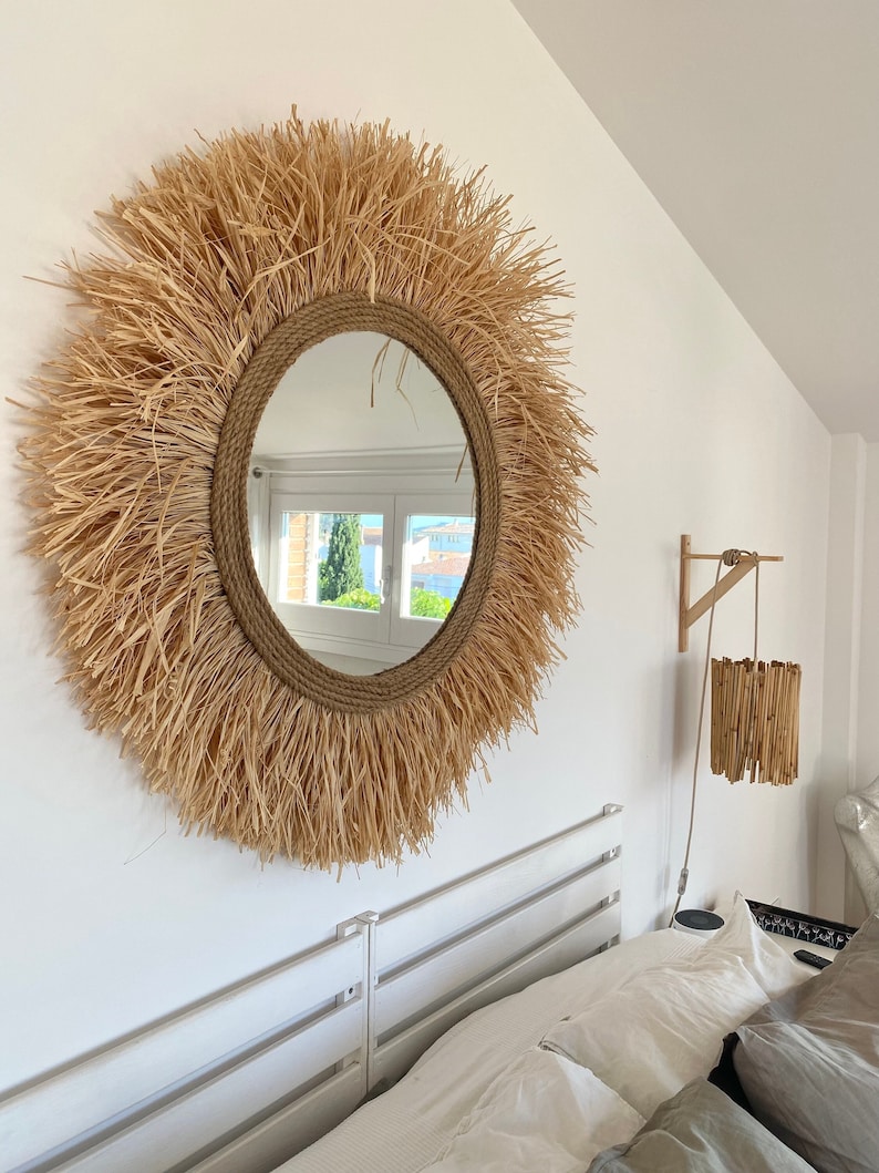 May include: A round mirror with a natural woven straw frame. The mirror is hanging on a white wall above a white bed frame.
