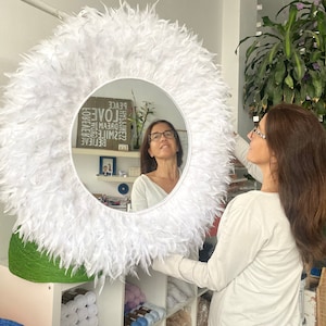 May include: A white feather mirror with a round frame. The mirror is reflecting a woman with brown hair wearing glasses. The mirror is hanging on a wall in a room with shelves filled with yarn.