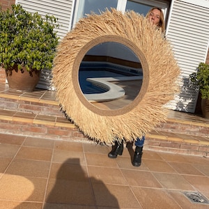 Pode incluir: Um espelho redondo grande com uma moldura tecida natural. A moldura &eacute; feita de um material semelhante &agrave; palha e tem uma franja ao redor da borda. O espelho reflete uma piscina e uma pessoa em p&eacute; na frente dele.