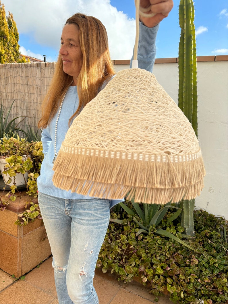May include: A beige woven lampshade with a fringe trim. The lampshade is being held by a person wearing a light blue sweater and blue jeans.