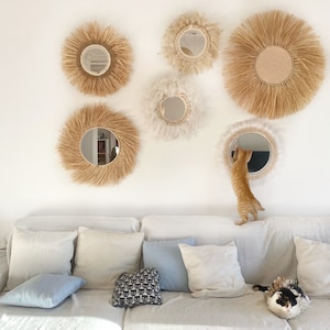 Pode incluir: Um sof&aacute; branco com almofadas e um gato sentado nele. A parede atr&aacute;s do sof&aacute; &eacute; decorada com v&aacute;rios espelhos redondos com molduras de fibra natural.