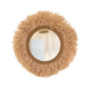 May include: A round mirror with a natural woven straw frame. The frame is thick and has a fringe of straw around the edge. The mirror is reflecting a room with a window and white curtains.