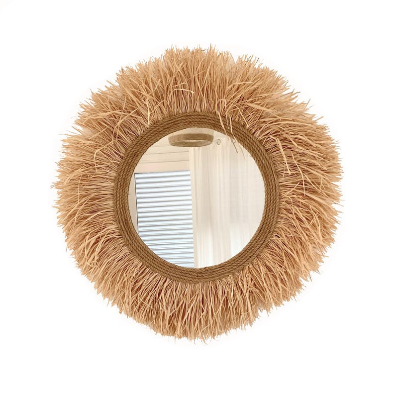 May include: A round mirror with a natural woven straw frame. The frame is thick and has a fringe of straw around the edge. The mirror is reflecting a room with a window and white curtains.