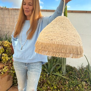 May include: A beige woven lampshade with a fringe trim. The lampshade is hanging from a white cord and is being held by a person.