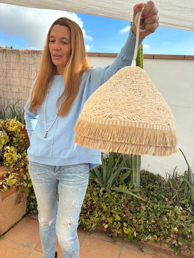 May include: A beige woven lampshade with a fringe trim. The lampshade is hanging from a white cord and is being held by a person.