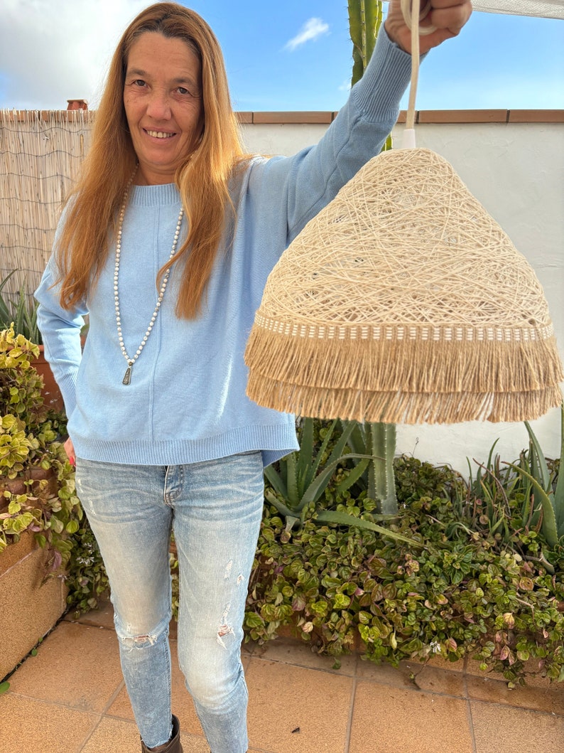 May include: A woman holds up a large, beige, woven pendant light with a fringe trim. The light is hanging from a white cord. The woman is wearing a light blue sweater and blue jeans.