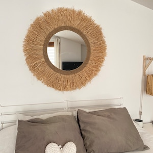 May include: A round mirror with a natural woven frame, hanging above a bed with grey linen pillows and a white heart-shaped pillow.
