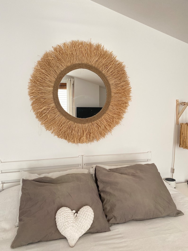May include: A round mirror with a natural woven frame, hanging above a bed with grey linen pillows and a white heart-shaped pillow.