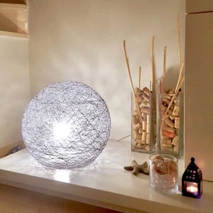 Peut inclure: Une lampe sphérique en corde grise avec une lumière blanche sur une étagère blanche. Il y a deux vases en verre remplis de bouchons de liège et un petit bol en verre avec des coquillages sur l'étagère.