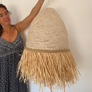 K&ouml;nnte beinhalten: Eine gro&szlig;e, beige, gewebte Pendelleuchte mit einem Fransen aus Naturfasern. Die Leuchte h&auml;ngt an einem wei&szlig;en Kabel.