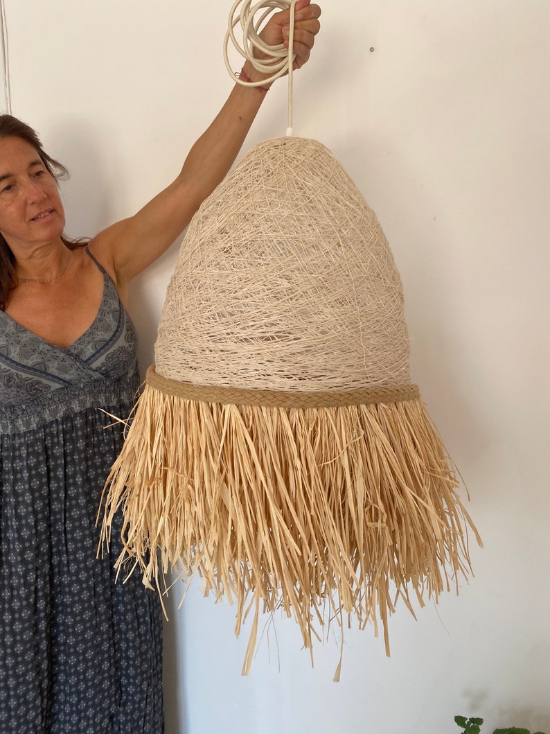 K&ouml;nnte beinhalten: Eine gro&szlig;e, beige, gewebte Pendelleuchte mit einem Fransen aus Naturfasern. Die Leuchte h&auml;ngt an einem wei&szlig;en Kabel.