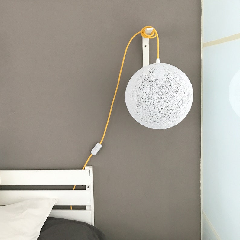 Peut inclure: Un luminaire suspendu rond en forme de corde blanche est suspendu &agrave; un cordon blanc et une fixation au plafond blanche. Le cordon est jaune et poss&egrave;de un interrupteur blanc. La lumi&egrave;re est suspendue dans une pi&egrave;ce avec une t&ecirc;te de lit en bois blanc.