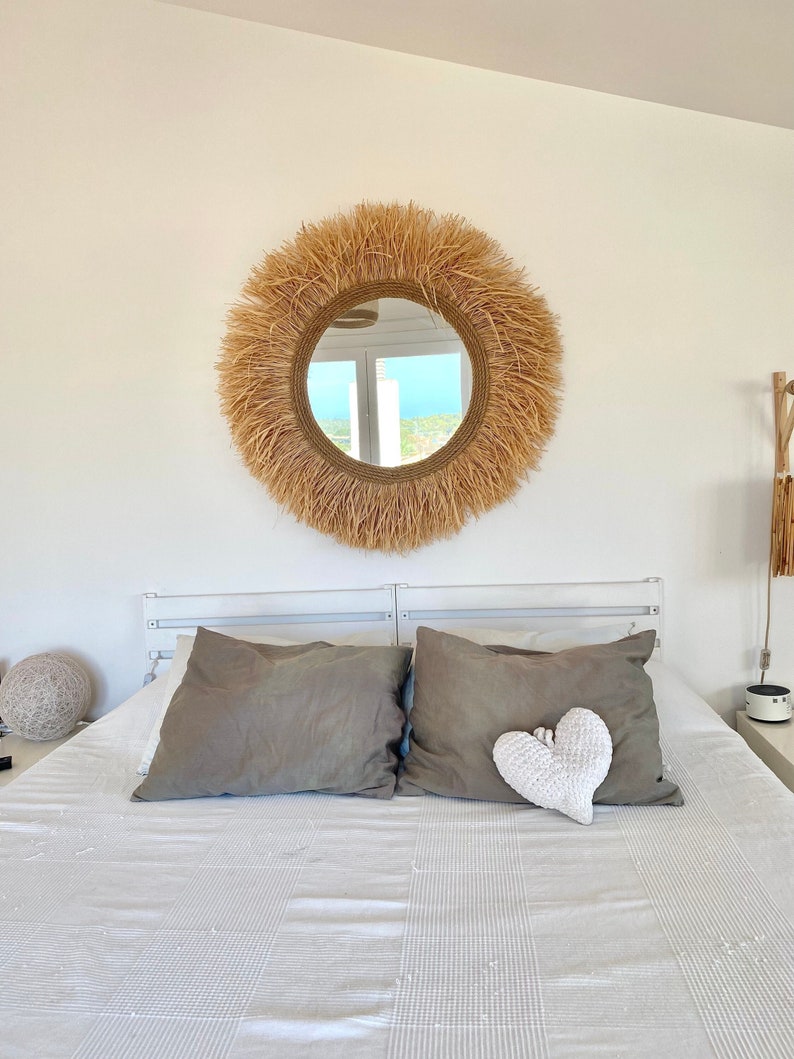 May include: A white bed with a grey headboard and two grey pillows. A white crocheted heart sits on the bed. A large round mirror with a woven straw frame hangs above the bed.