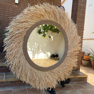 Pode incluir: Um grande espelho redondo com uma moldura de palha natural tecida. O espelho est&aacute; pendurado em uma parede de tijolo e reflete um jardim com uma pequena &aacute;rvore e um c&atilde;o.