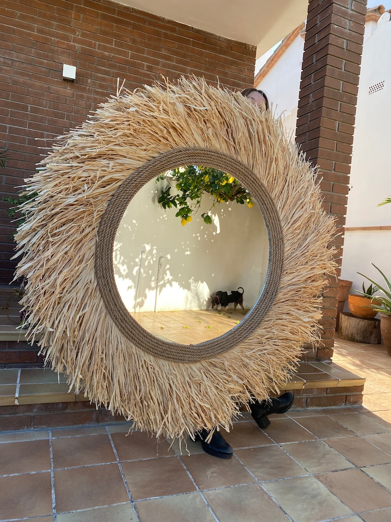 Pode incluir: Um grande espelho redondo com uma moldura de palha natural tecida. O espelho est&aacute; pendurado em uma parede de tijolo e reflete um jardim com uma pequena &aacute;rvore e um c&atilde;o.