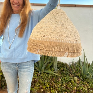 May include: A beige woven lampshade with a fringe trim. The lampshade is hanging from a white cord.