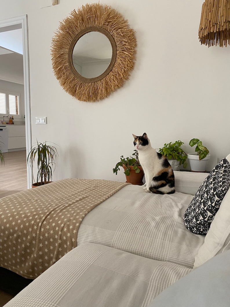 May include: A white and black cat sits on a bed with a tan and white striped blanket and a white and grey striped pillow. A large round mirror with a natural fibre frame hangs on the wall behind the bed.