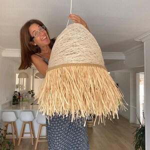 K&ouml;nnte beinhalten: Eine Frau h&auml;lt eine gro&szlig;e, runde, beige Lampenschirm mit einer Fransen aus Naturfasern hoch. Der Lampenschirm h&auml;ngt an einem wei&szlig;en Kabel.