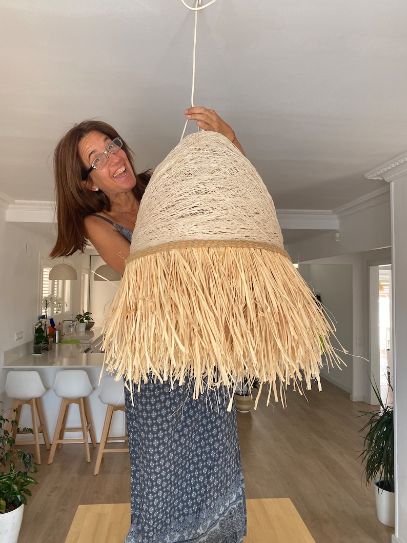 K&ouml;nnte beinhalten: Eine Frau h&auml;lt eine gro&szlig;e, runde, beige Lampenschirm mit einer Fransen aus Naturfasern hoch. Der Lampenschirm h&auml;ngt an einem wei&szlig;en Kabel.