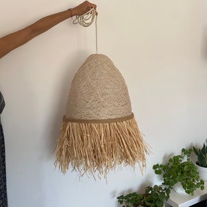 K&ouml;nnte beinhalten: Eine beige gewebte Pendelleuchte mit Fransenbesatz. Die Leuchte h&auml;ngt an einem wei&szlig;en Kabel.