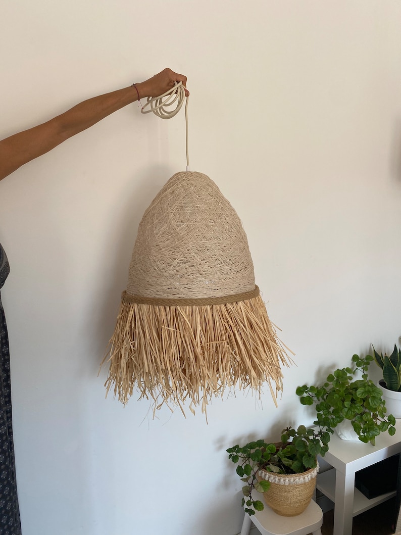 K&ouml;nnte beinhalten: Eine beige gewebte Pendelleuchte mit Fransenbesatz. Die Leuchte h&auml;ngt an einem wei&szlig;en Kabel.