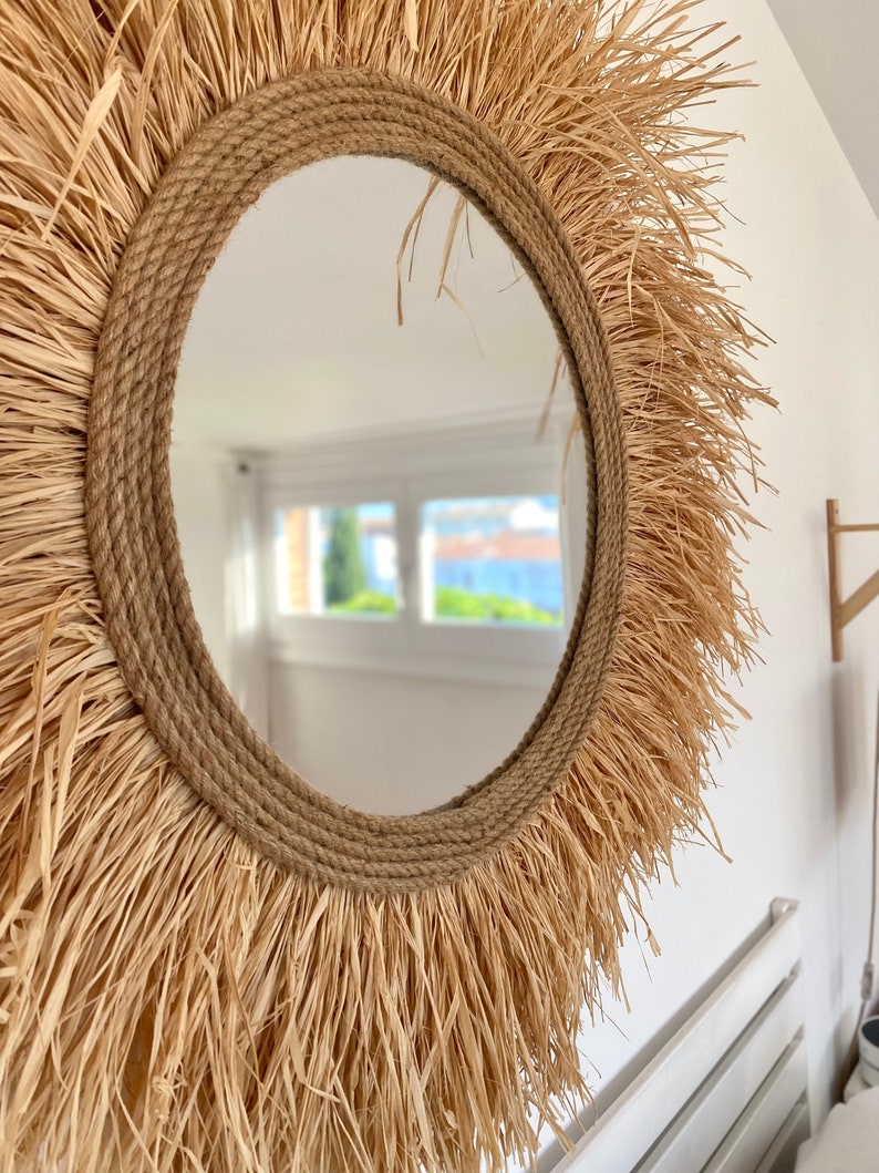 May include: A round mirror with a natural woven frame. The frame is made of straw-like material and has a rope border. The mirror is reflecting a room with a window.
