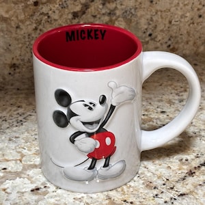 K&ouml;nnte beinhalten: Wei&szlig;er Keramikbecher mit roter Innenseite und wei&szlig;em Henkel. Der Becher zeigt ein erhabenes Bild von Mickey Mouse in seinem klassischen Outfit. Das Wort "MICKEY" ist schwarz auf dem Rand gedruckt.