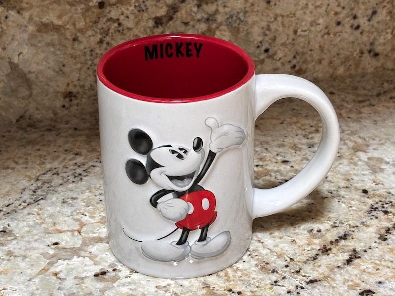 K&ouml;nnte beinhalten: Wei&szlig;er Keramikbecher mit roter Innenseite und wei&szlig;em Henkel. Der Becher zeigt ein erhabenes Bild von Mickey Mouse in seinem klassischen Outfit. Das Wort "MICKEY" ist schwarz auf dem Rand gedruckt.