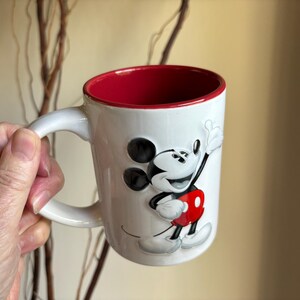 K&ouml;nnte beinhalten: Wei&szlig;er Keramikbecher mit rotem Innenraum und Henkel. Der Becher zeigt ein erhabenes Bild von Mickey Mouse in seinem klassischen Outfit. Das Design umfasst schwarze Ohren, eine rote Hose und wei&szlig;e Handschuhe. Ein lustiges und farbenfrohes Sammlerst&uuml;ck.