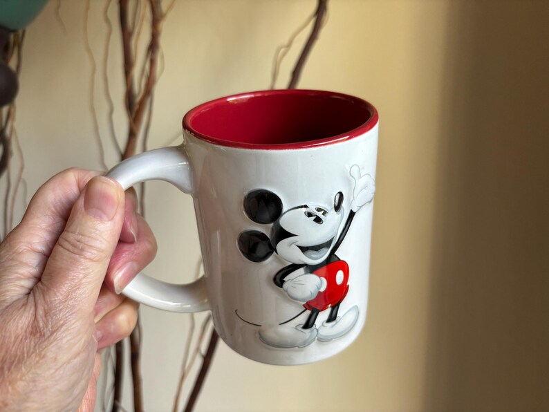 K&ouml;nnte beinhalten: Wei&szlig;er Keramikbecher mit rotem Innenraum und Henkel. Der Becher zeigt ein erhabenes Bild von Mickey Mouse in seinem klassischen Outfit. Das Design umfasst schwarze Ohren, eine rote Hose und wei&szlig;e Handschuhe. Ein lustiges und farbenfrohes Sammlerst&uuml;ck.
